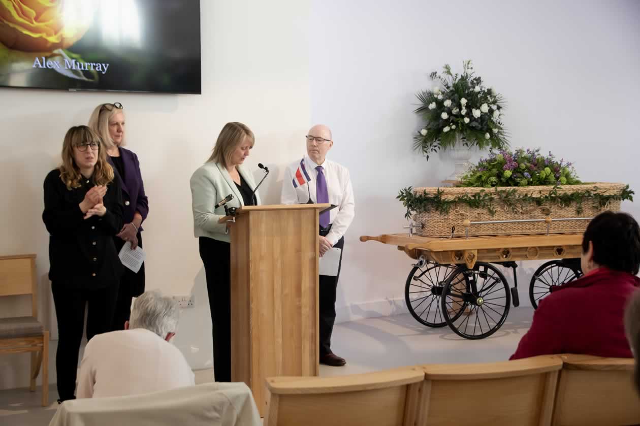 Staged funeral celebrants talk at a lectern beside a mock coffin