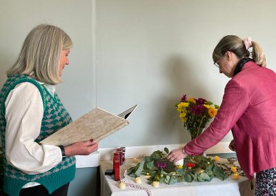A female celebrant with a folder instructs a woman to place a flower in the water for a flower ceremony